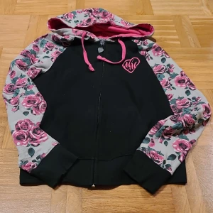 Blommig hoodie från Metal Mulisha - Snygg hoodie från Metal Mulisha med svart kropp och grå ärmar och huva täckta av rosa blommor. Rosa snören och broderad logga på bröstet. Dragkedja framtill och mjukt material som känns skönt mot huden. Perfekt för dig som gillar streetstyle och unika detaljer.