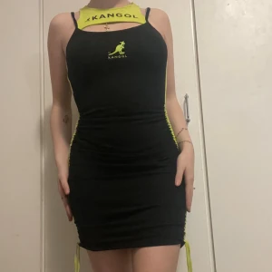 Svart och limegrön tight klänning Kangol - Supersnygg svart kortklänning från Kangol med limegröna detaljer och logga framtill. Klänningen har smala axelband, tight passform och cool snörning i sidorna. Materialet är stretchigt och känns sportigt. Perfekt för dig som vill sticka ut med en unik look!