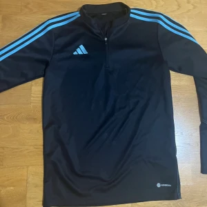 Svart Adidas half zip tröja S - Svart långärmad tröja från Adidas med ljusblå ränder längs axlarna och half zip framtill. Tröjan har Aeroready-tryck nere vid höften och är gjord i ett mjukt syntetmaterial som passar perfekt till träning. Klassisk Adidas-logga på bröstet.