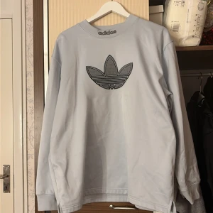 Ljusgrå sweatshirt från Adidas - Säljer en ljusgrå sweatshirt från Adidas med stort broderat trefoil-logo i svart på bröstet och Adidas-text på kragen. Tröjan har långärmad passform och är gjord i mjuk bomullsmix. Perfekt för dig som gillar streetwear och sportig stil.