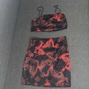 Svart och röd bandeau topp+kjol med drakmönster - Säljer en snygg bandeau topp med tunna axelband+kjol i svart med röda drakar som mönster. Toppens material är mjukt och stretchigt, troligen polyester, och har en croppad passform som passar perfekt till kjol eller jeans. Perfekt för dig som gillar asiatiska influenser och coola prints.