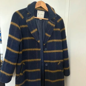 Randigt ullkappa från R Éssentiel - Mörkblå kappa i ullmix med breda horisontella senapsgula ränder. Klassisk krage, knäppning med två knappar och fickor fram. Fodrad insida för extra komfort. Perfekt för dig som vill sticka ut med en snygg och mysig jacka.