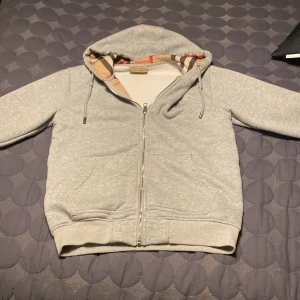 Grå Burberry hoodie med dragkedja - Snygg grå hoodie från Burberry med klassiskt rutigt foder i huvan. Tröjan har hel dragkedja, två fickor fram och justerbar huva med snören. Perfekt för dig som gillar stilrena och exklusiva plagg med en twist.