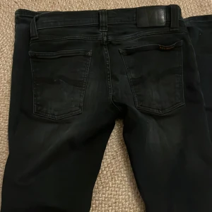 Nudie jeans - Säljer ett par svarta jeans från Nudie Jeans i modellen skinny med smal passform och klassiska fem fickor. Jeansen har svarta sömmar och en diskret logga på bakfickan. Perfekt för dig som gillar en stilren och tajt look. Passar mig som är 175 väger 57kg.