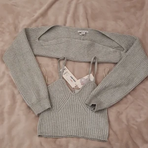 Grå ribbad bolerotopp från New Yorker  - Säljer en snygg grå ribbad bolerotopp från New Yorker. Toppen består av en croppad ärmlös topp med smala axelband och en matchande bolero med långa ärmar. Perfekt för lager-på-lager-look och riktigt trendig vibe.