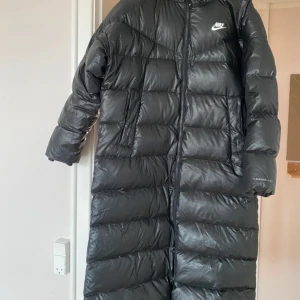Svart lång dunjacka från Nike - Säljer en svart lång dunjacka från Nike med huva och Therma-FIT teknologi. Jackan har en clean look med diskret logga på bröstet, dragkedja framtill och två sidofickor. Perfekt för kalla dagar när du vill hålla värmen och se fresh ut.