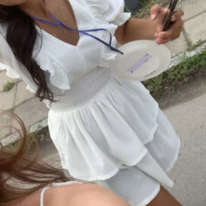 Vit volangklänning från Gina Tricot - Supersöt vit kortklänning från Gina Tricot med v-ringning, volanger på axlarna och puffärmar. Klänningen har smock i midjan och är tillverkad i ett lätt, krinklat bomullstyg som ger en luftig känsla. Perfekt för sommarens alla tillfällen! Använt endast en gång och klänningen är som ny! 