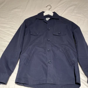 Mörkblå overshirt från John Henric XS - Snygg mörkblå overshirt från John Henric i bomull. Jackan har klassisk krage, fyra fickor framtill och stängs med knappar. Perfekt lager-på-lager-plagg med stilren och enkel design.