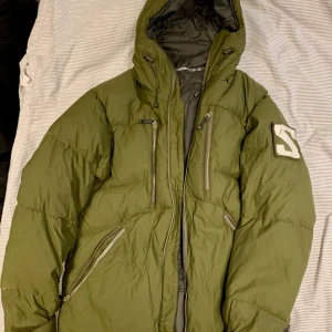 Grön Alpin dunjacka från Salomon - Säljer en grön dunjacka från Salomon med huva, insydd fleecehuva, utanpåliggande dunhuvs samt flera dragkedjeförsedda fickor. Jackan har ett stort S-märke på vänster ärm och quiltad design. Insidan är grå med tryckt motiv och praktiska innerfickor. Perfekt för kalla dagar. Har avtagbart snölås och midjedel.