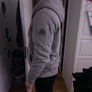 Grå ”hoodie” jacka från Moose Knuckles -  huva och svarta snören. Jackan har en snygg logotyp på ärmen, ribbade muddar och dragkedja framtill. Perfekt för dig som vill ha en stilren och bekväm jacka. Modellen är 188cm 