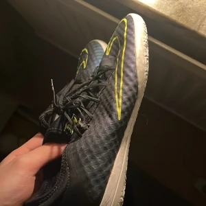 Nike svarta fotbollsskor med neon - Säljer ett par svarta Nike fotbollsskor med neon-gula detaljer och snörning. Skorna har en rund tå och en platt sula med mönster i blått och gult undertill för extra grepp. Ovandelen är i syntetmaterial med en sportig look och tydlig Nike-logga på sidan.