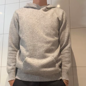 Grå stickad hoodie med huva - Mysig grå stickad hoodie med huva och känguruficka framtill. Tröjan har lång ärm och ribbade muddar vid ärmslut och nederkant. Perfekt för kyliga dagar när du vill vara både bekväm och snygg.