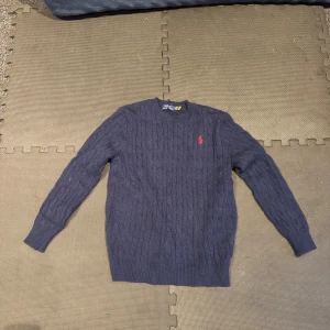 Marinblå stickad tröja från Polo Ralph Lauren - Snygg marinblå stickad tröja från Polo Ralph Lauren med klassiskt kabelstickat mönster och röd broderad logga på bröstet. Rund hals och långa ärmar med ribbade muddar. Storlek M, passar folk ca 170-180cm