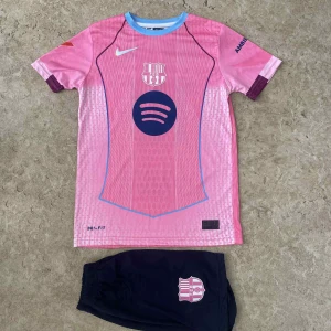Barça Lamine Yamal rosa fotbollströja - Säljer en FC Barcelona fotbollströja med Lamine Yamal och nummer 10 på ryggen. Tröjan är rosa med blå detaljer, har korta ärmar och är gjord i Dri-Fit material. Fram finns Spotify-logga och klubbmärke, bak UNHCR-tryck. Matchande mörkblå shorts med klubbmärke ingår.