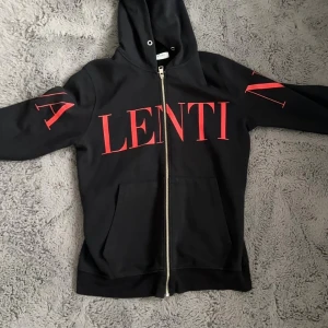 Svart hoodie från Valentino med röd print - Svart hoodie från Valentino med stor röd logoprint över bröstet och armarna. Tröjan har huva, dragkedja framtill och två fickor. Materialet är mjuk bomull och passformen är normal. Perfekt för dig som gillar exklusiva streetwear-vibbar.