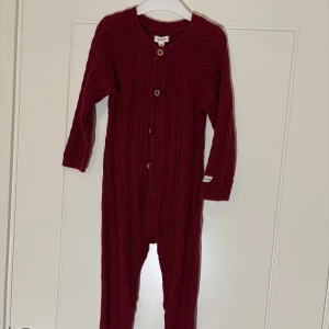 Newbie - stickad overall, storlek 86 - Vinröd stickad overall från Newbie med snyggt kabelmönster och träknappar framtill. Mjuk och varm, perfekt för kyliga dagar. Långa ärmar och ben, ribbade muddar och rund hals.  Skickar med frakt eller avhämtning i Växjö. Vid flera köp kan du som köpare välja samfrakt. Fraktpriset ingår inte i priset. 