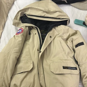 Beige dunjacka Canada Goose XS - Säljer en beige dunjacka från Canada Goose i storlek XS. Jackan har huva, flera fickor med lock och dragkedja, samt den klassiska röda Canada Goose Arctic Program-patchen på ärmen. Perfekt för kalla dagar och har en clean, stilren look. Priset går absolut och diskuteras 