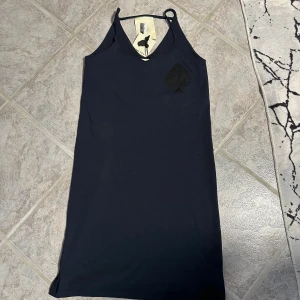 Marinblå klänning från 5 Preview - Marinblå kortklänning från 5 Preview med smala axelband och v-ringning. Klänningen har en stor, diskret spaderdetalj på bröstet och är tillverkad i mjuk bomull. Enkel och stilren design som känns modern och cool.