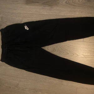 Svarta mjukisbyxor från Nike - Svarta mjukisbyxor från Nike med vit logga på vänster lår. Byxorna har elastisk midja med snörning och smal passform nedtill. Perfekta för chill eller träning, tillverkade i mjukt bomullsmaterial för extra komfort.