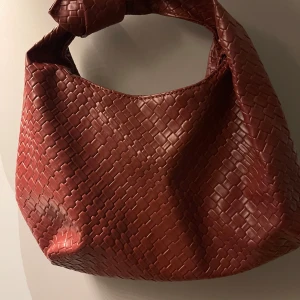 Flätad röd axelväska i skinn - Säljer en snygg brun axelväska med flätad design och knut på handtaget. Väskan är tillverkad i skinn och har en mjuk, slouchy form som ger en cool och trendig vibe. Perfekt för dig som vill ha en unik accessoar med stil.