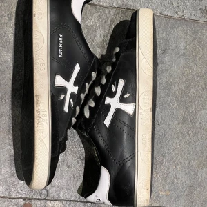 Svarta Premiata Steven - Snygga svarta sneakers från Premiata med vita detaljer och logga på sidan. Skorna har vit sula med tryck, grå snörning och svart ovandel i skinn och mocka. Bakpartiet är vitt med Premiata-tryck. Perfekta för dig som gillar stilrena och trendiga sneakers.