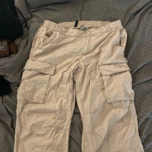 Beiga cargopants från H&M Divided - Säljer ett par beiga cargopants från H&M Divided i storlek 50. Byxorna har breda ben, elastisk midja och stora fickor på sidorna. de har en avslappnad, loose passform som är perfekt för en chill streetwear-look.