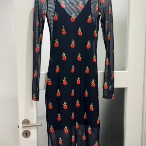 Svart meshklänning med eldprint - Svart lång meshklänning med transparenta långa ärmar och coolt eldprint i rött och gult över hela klänningen. Klänningen har rund hals och figurnära passform, perfekt för dig som vill sticka ut med en edgy vibe.