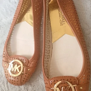 Bruna ballerinas från Michael Kors - Snygga bruna ballerinas från Michael Kors i skinn med perforerat mönster och rund tå. Dekorerade med ett stort guldfärgat MK-spänne framtill. Perfekta för dig som vill ha en klassisk men trendig look med lyxig känsla.