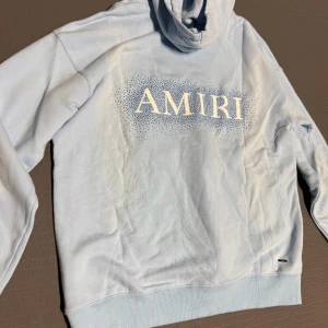 Ljusblå AMIRI hoodie  - Snygg ljusblå hoodie från AMIRI med stort vitt logoprint på bröstet och mindre logga på framsidan. Tröjan har huva, magficka och ribbade muddar. Materialet är mjuk bomull och passformen är normal. Perfekt för dig som gillar streetwear och vill sticka ut.