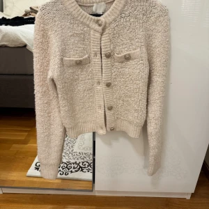 Beige kofta från H&M med knappar - Säljer en beige kofta från H&M i storlek XS. Koftan har en fluffig, stickad struktur och ribbade muddar. Den har långa ärmar, två dekorativa bröstfickor med knappar och knäppning framtill. Perfekt för dig som gillar mysiga och trendiga plagg.