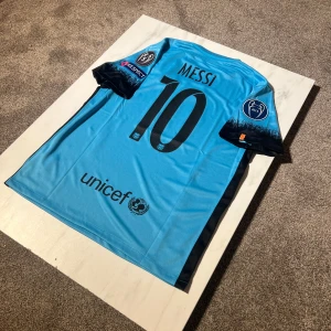 Barça Messi bortatröja Nike 2015/16 - Säljer en ikonisk FC Barcelona bortatröja från säsongen 2015/16 med Messi 10 på ryggen. Tröjan är ljusblå med svarta detaljer, korta ärmar och broderade klubbmärken, FIFA-märke och Champions League-patchar. Materialet är lätt och andas, perfekt för fotboll eller samling.