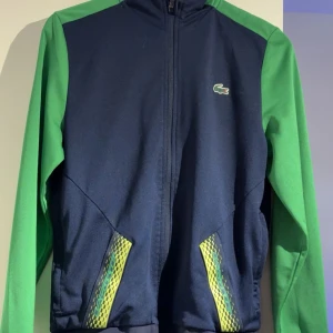 Blå och grön zip tröja från Lacoste - Sportig zip tröja från Lacoste med marinblå kropp och gröna ärmar. Tröjan har dragkedja framtill, långärmad design och coola gula detaljer vid fickorna. Klassisk Lacoste-logga på bröstet och bekväm passform. Perfekt för dig som gillar streetwear och sportig stil.