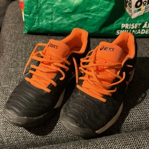 Svarta och orangea Asics sneakers - Säljer ett par svarta och orangea Asics sneakers med snygg sportig design. Skorna har snörning, platt sula och är tillverkade i syntetmaterial. Perfekta för dig som gillar att sticka ut med färg och vill ha bekväma skor till träning eller vardag. Använd bara ett fåtal tennisträningar men underbart sköna.