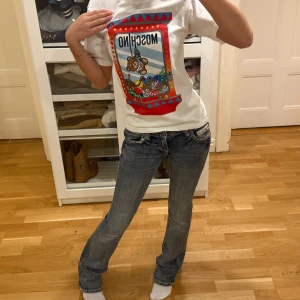Vit Moschino t-shirt med nalletryck - Vit t-shirt från Moschino med färgglatt tryck av en nallemaskin och flera nallar på bröstet. Köpt på NK. Klassisk rund hals och korta ärmar. Skön passform och tillverkad i mjuk bomull. Perfekt statement-plagg för dig som gillar lekfull stil. 