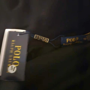 Svart hoodie från Polo Ralph Lauren - Svart hoodie från Polo Ralph Lauren med dragkedja och huva. Klassisk design med snörning i huvan och röd broderad logga på bröstet. Tillverkad i mjuk bomull, perfekt för chill dagar. Långärmad och med ribbade muddar för extra komfort.