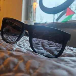 Svarta Ray-Ban Justin solglasögon - Säljer ett par svarta Ray-Ban Justin solglasögon med breda bågar och rektangulär form. Modellen är RB4165 och de är tillverkade i Italien. Snygg och klassisk design som passar perfekt till sommaren. Linsfärgen är mörk och bågarna är i matt plast.