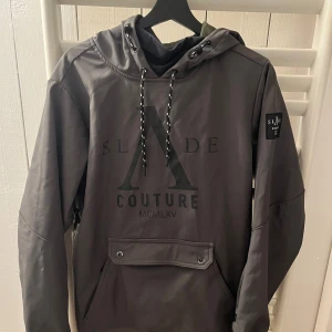 Mörkgrön skidjacka  från Slade Couture - Snygg Mörkgrön skidjacka  från Slade Couture med stor logga på bröstet och patch på ärmen. Jackan  har  huva med snörning, praktisk magficka med knappdetaljer och dragkedjor på ärmarna. Perfekt för dig som gillar streetwear och vill ha en unik look. Hör av er vid frågor 