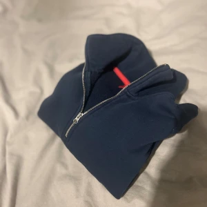 Marinblå half zip tröja från RYVLS - Marinblå långärmad tröja från RYVLS med half zip och hög krage. Tröjan har ribbade muddar vid ärmslut och nederkant samt en snygg röd detalj på insidan av kragen. Perfekt för dig som gillar enkel och stilren design.