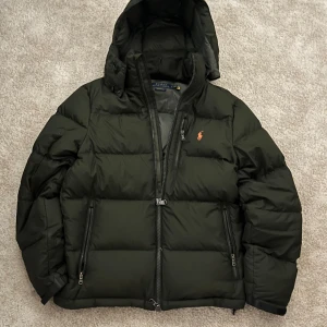 Mörkgrön pufferjacka från Polo Ralph Lauren - Snygg mörkgrön pufferjacka från Polo Ralph Lauren med huva och orange logga på bröstet. Jackan har flera dragkedjeförsedda fickor och quiltad design. Perfekt för kalla dagar och har en clean, sportig look.
