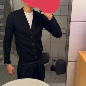 Svart kofta från Dressman, långärmad - Svart kofta från Dressman med v-ringad hals och fyra knappar framtill. Långärmad och ribbad vid muddar och nederkant. Mjuk och skön, perfekt för lager-på-lager. Passar dig som gillar enkel och stilren look. Modell 175cm