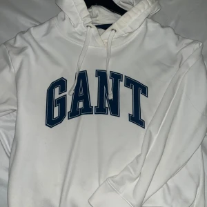 Vit hoodie från GANT med blå logga - Vit hoodie från GANT med stor blå logotyp framtill. Tröjan har huva med snörning och långa ärmar. Mjuk och skön bomullskvalitet som passar perfekt när du vill ha en chill och stilren look. Klassisk passform och ribbade muddar vid ärmslut och nederkant.