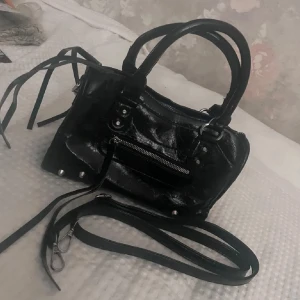 Svart handväska med dragkedja och nitar - Cool svart handväska i glansigt skinnliknande material med silverfärgade nitar och detaljer. Väskan har både handtag och en avtagbar axelrem, samt flera dragkedjor och dekorativa remmar. Insidan har ett fack med dragkedja för småsaker.