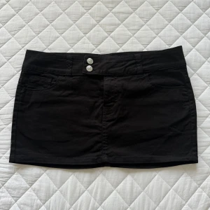 Svart kortkjol från H&M Divided - Svart kortkjol från H&M Divided med två silvriga knappar framtill, klassiska jeansfickor både fram och bak samt bälteshällor. Kjolen är gjord i bomull med lite elastan för skön stretch. Perfekt för dig som gillar enkel och clean stil.