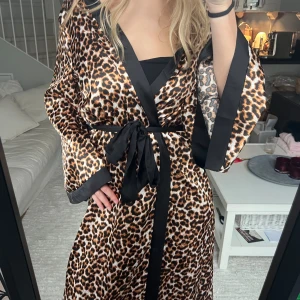 Leopardmönstrad morgonrock i satin - Snygg satinmorgonrock med leopardmönster och svarta satinliknande detaljer längs kanterna. Den har vida ärmar och knyts i midjan med ett brett svart band. Älskar denna, så snygg och mysig! Hör av dig till mig för fler bilder 🫶🏼