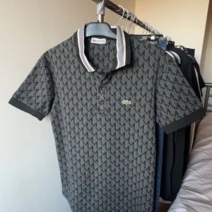 Grå mönstrad Lacoste piké  - BYTE FUNKAR! Snygg grå t-shirt från Lacoste med diskret geometriskt mönster över hela plagget. Klassisk krage med vita och svarta ränder, korta ärmar och knappar framtill. Ikoniska Lacoste-loggan på bröstet. Perfekt för dig som gillar stilren design. Har använt den 2 gånger