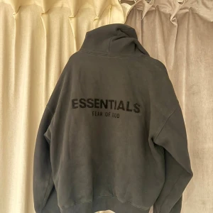Svart Essentials Fear of God hoodie - Svart hoodie från Essentials Fear of God med stor logga på ryggen. Oversized passform och huva. Tillverkad i mjuk bomull och polyesterblandning, perfekt för chill dagar. Klassisk look med nedhasade axlar och breda ärmar.