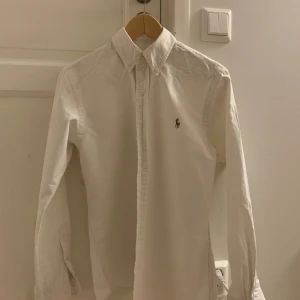 Vit slim fit skjorta från Ralph Lauren - Vit skjorta från Ralph Lauren i slim fit-modell, tillverkad i mjuk bomull. Klassisk design med knappar framtill, button-down krage och broderad logga på bröstet. Långärmad och stilren, perfekt för dig som gillar clean och tidlös look.