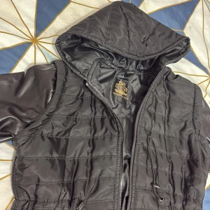 Svart lång pufferjacka med huva - Svart lång pufferjacka från Luxury Polo med quiltad kropp och släta skinnärmar. Jackan har huva, dragkedja framtill och snörning i midjan för extra passform. Perfekt för kalla och regniga dagar , modern look.