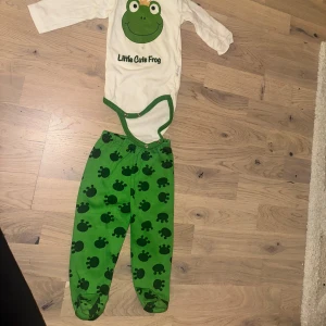 Grodset med Body, Byxor och Mössa - Säljer ett par gröna byxor med mörkgröna grodmönster, perfekta för små barn. Byxorna har resår i midjan och är gjorda i mjukt bomullsmaterial. Passar dig som gillar roliga prints och färgglada kläder till de minsta.