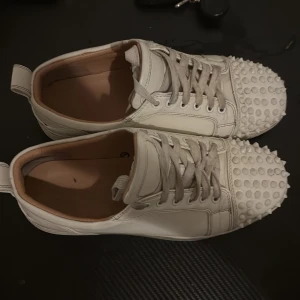 Vita Christian Louboutin sneakers med nitar - Säljer ett par vita sneakers från Christian Louboutin i skinn med ikoniska röda sulor. Skorna har snörning och coola nitar över tån för en edgy vibe. Låg modell med rund tå och klassisk siluett. Perfekta för dig som vill sticka ut med lyxig streetstyle.jätte bekväma !! Tveka inte på att höra av dig vid intresse